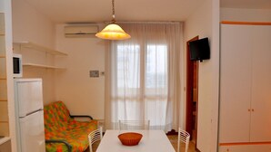 A compact living space with a kitchenette, a dining area with a table and chairs, a colorful sofa, and a television mounted on the wall. - Relaxing studio apartment with side sea view (Bibione)