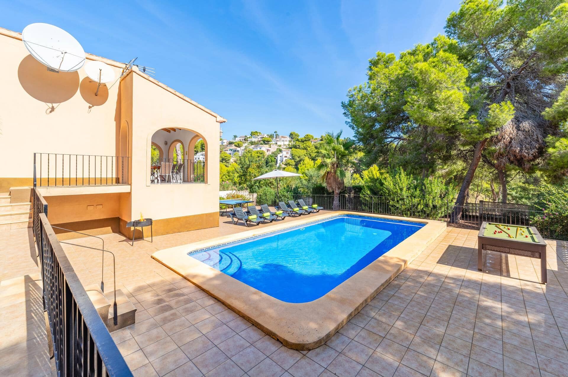 A swimming pool with a tiled deck, a covered seating area with chairs, and a view of a residential area with trees and houses.