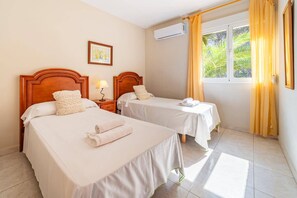 A hotel room with two beds, a nightstand, an air conditioning unit, a window with curtains, and a framed picture on the wall.