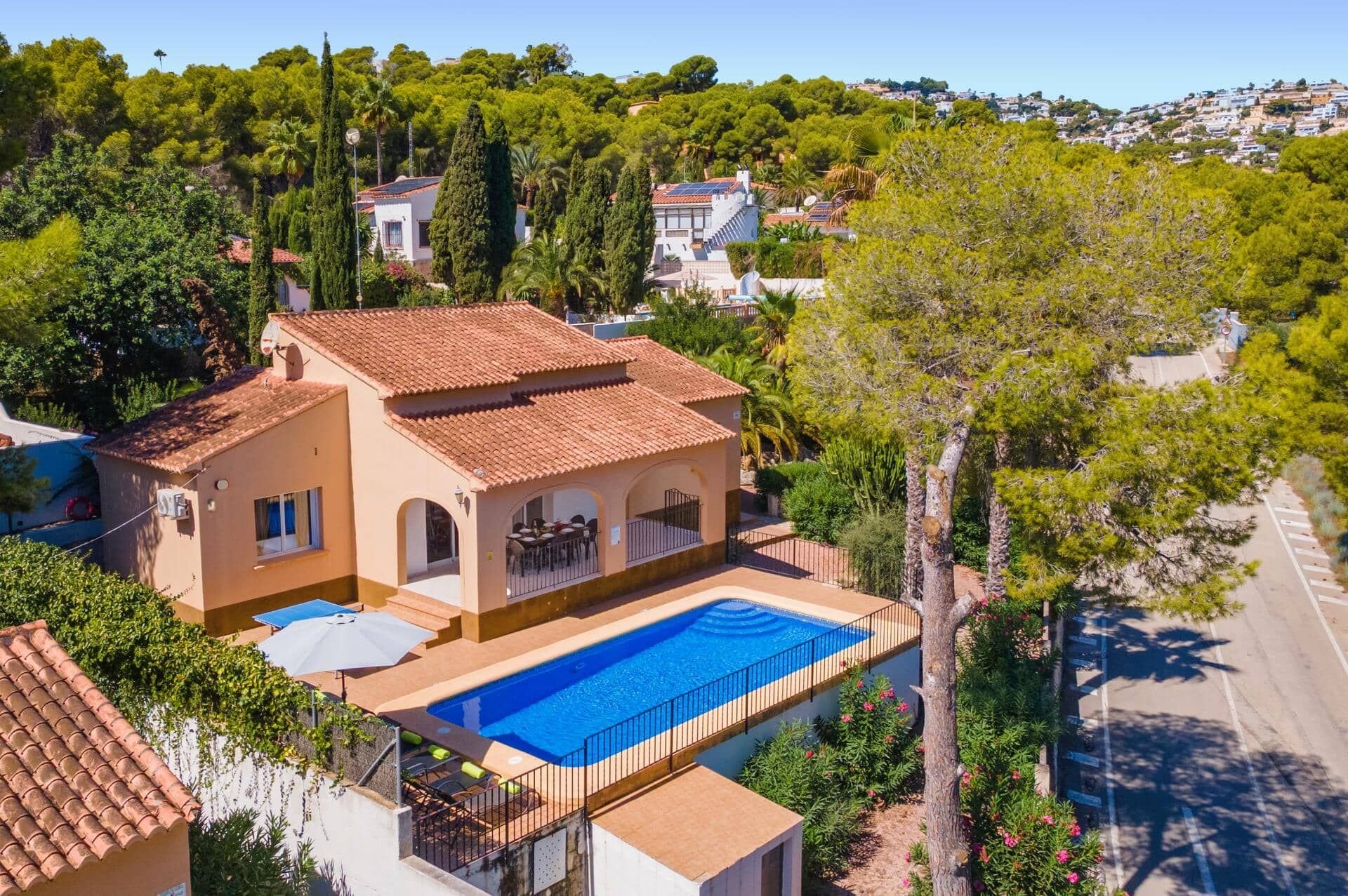Una villa de estilo mediterráneo con piscina, rodeada de exuberante vegetación y un cielo azul despejado.