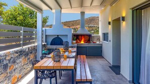 An outdoor patio with a stone wall, a wooden table with benches, a grill, and a view of the hills. - Villa Manefic by Villa Plus (Malevizi)