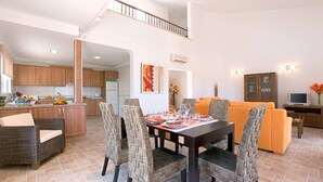 A spacious living area with a dining table set for four, surrounded by wicker chairs. A kitchen is visible in the background, and a staircase leads to an upper level. - Villa Tia Elanor by Villa Plus (Jávea)
