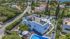 A modern white building with a rooftop pool, surrounded by greenery and a clear view of the sea.
