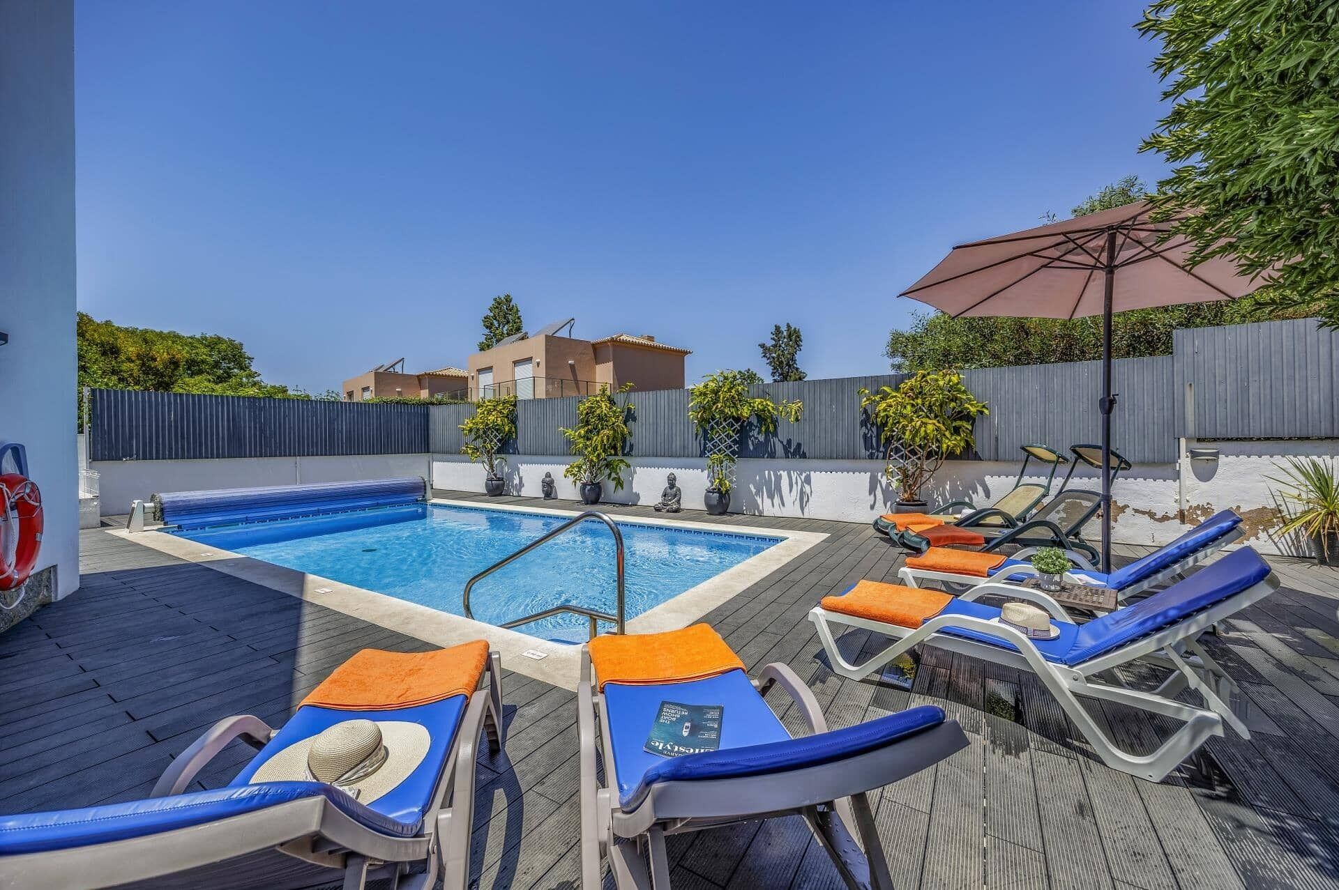 Piscina con sillones de jardín y sombrillas, rodeada por una terraza de madera y un muro bajo con plantas en macetas.