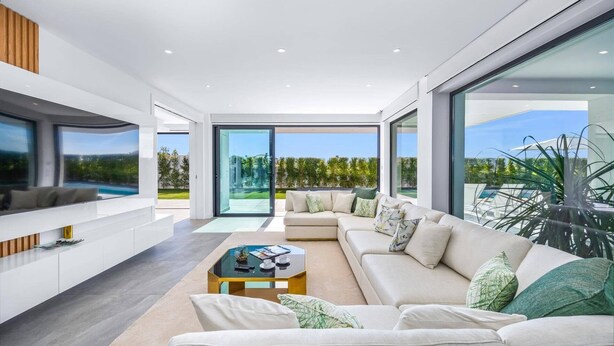 Ein modernes Wohnzimmer mit einem großzügigen Ecksofa, einem Glastisch und einem Fernseher auf einer weißen Konsole. Große Fenster bieten Blick auf einen Pool und viel Grün.