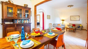 A dining area with a wooden table set for four, surrounded by orange chairs. A wooden cabinet with glass doors is to the left, and a living room with a sofa and a lamp is in the background.