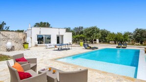 Una villa moderna con piscina, mobiliario exterior y un muro de piedra.