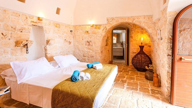 Una habitación de hotel con pared de piedra, una cama con ropa blanca, una puerta de madera y un jarrón con flores.