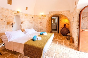 Una habitación de hotel con pared de piedra, una cama con ropa blanca, una puerta de madera y un jarrón con flores.