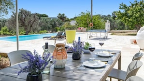 Un área de comedor al aire libre con una piscina al fondo, rodeada de follaje y un cielo despejado.