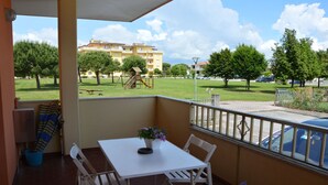 Un balcon avec une table et des chaises blanches, un pot de fleurs et une vue sur un terrain de jeux et des bâtiments au loin.