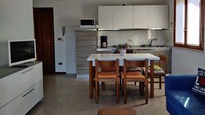 Une cuisine compacte avec des meubles blancs, un four à micro-ondes et une machine à café. On y trouve une table à manger avec quatre chaises et un canapé bleu avec un coussin.