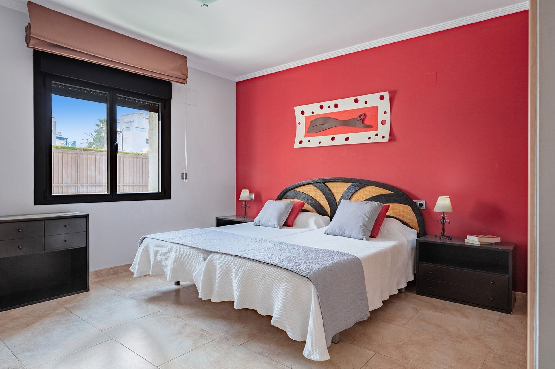 Un dormitorio con cama, mesitas de noche, una ventana con persianas y una pared de acento roja.