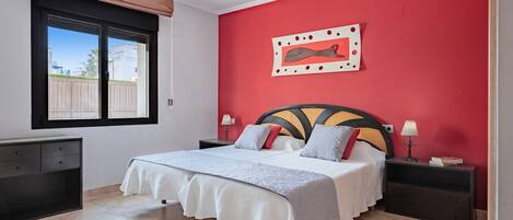 Un dormitorio con cama, mesitas de noche, una ventana con persianas y una pared de acento roja.