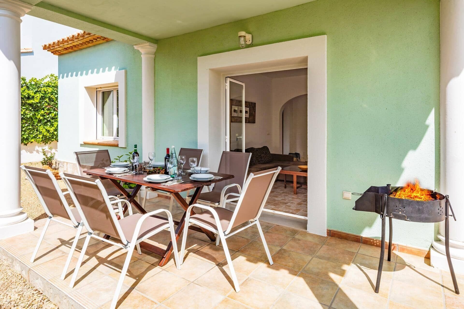 An outdoor dining area with a table set for four, surrounded by chairs, and a barbecue grill to the side.