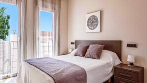A bedroom with a bed, a nightstand, a lamp, a wall-mounted artwork, and a view of the city through a window. - Villa Pomera Tres by Villa Plus (Beniarbeig)