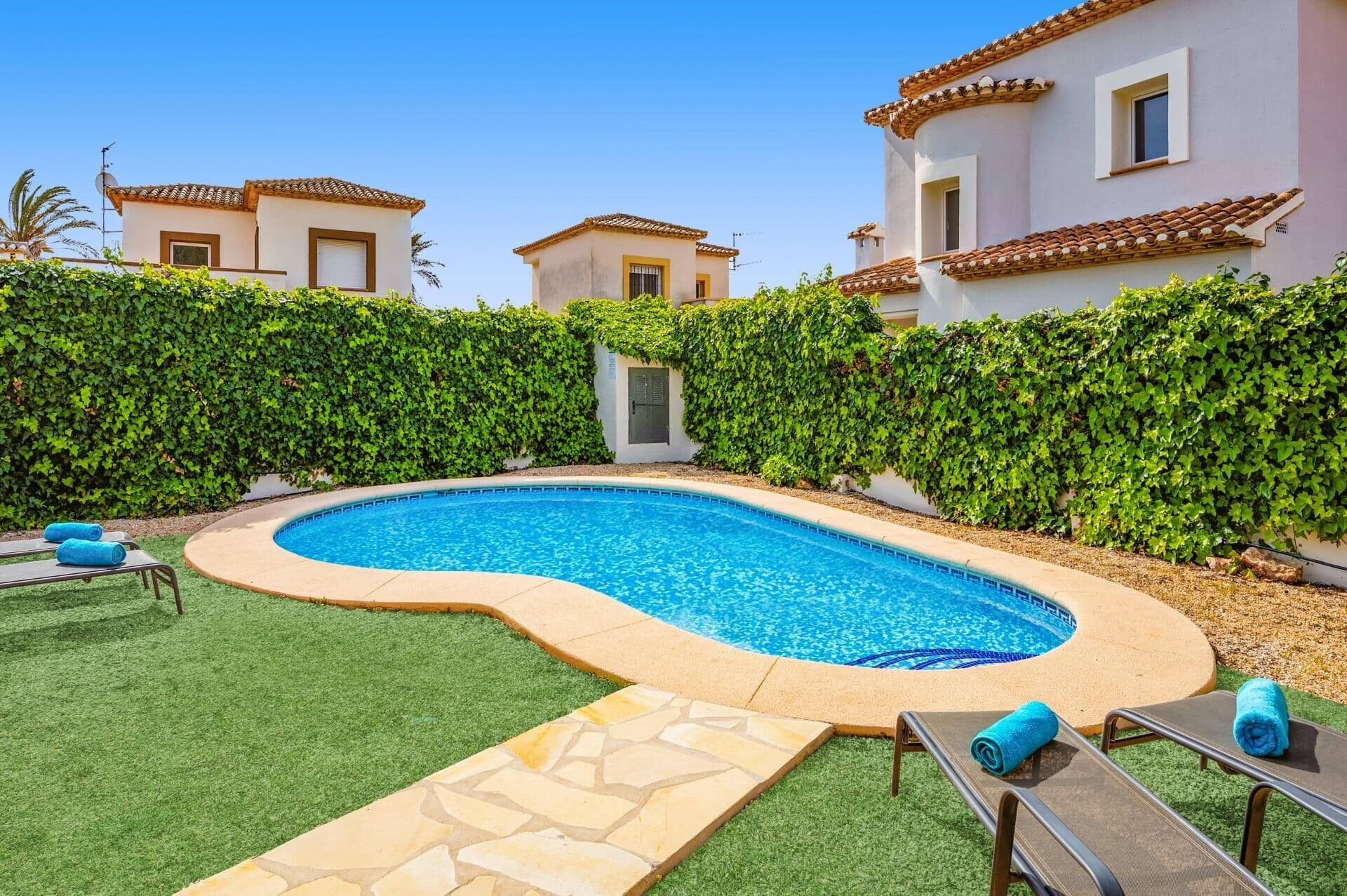 A swimming pool with a stone border, surrounded by artificial grass and a hedge.