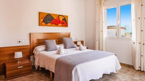 A hotel room with a wooden headboard, a large bed with grey and white bedding, a bedside table with a lamp, and a painting above the bed. - Villa Jazmin Blanca by Villa Plus (Beniarbeig)