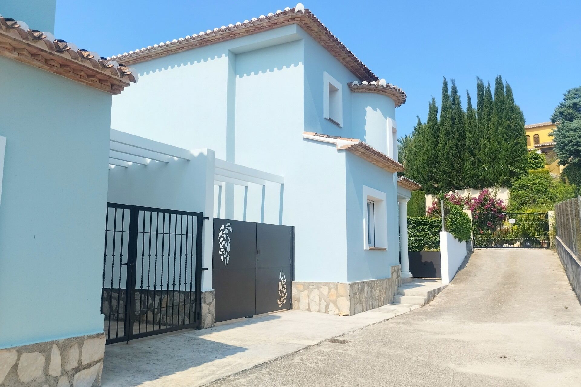 A light blue building with a tiled roof, a gated entrance, and a narrow street leading to it.