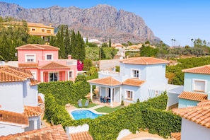 A residential area with houses featuring terracotta roofs, a swimming pool, and a mountainous backdrop.