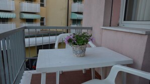 Un balcón con una mesa y sillas blancas, una cesta de mimbre con flores y una ventana.