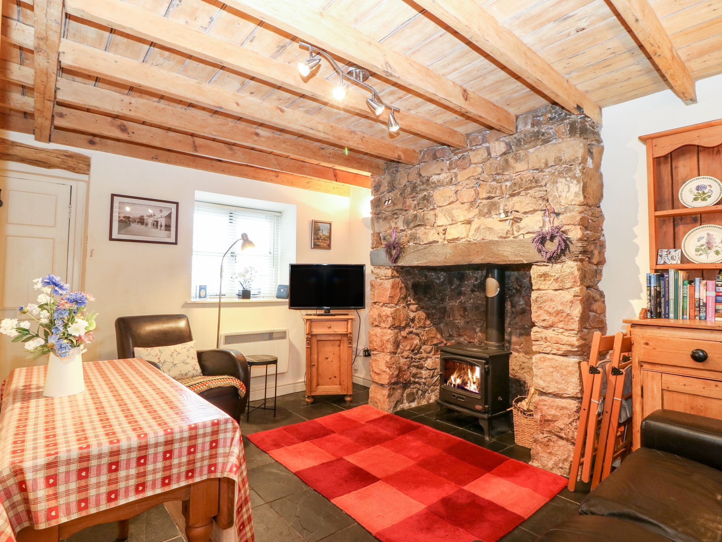 A cozy living room with a stone fireplace, wooden beams, a television, and a dining area with a checkered tablecloth.