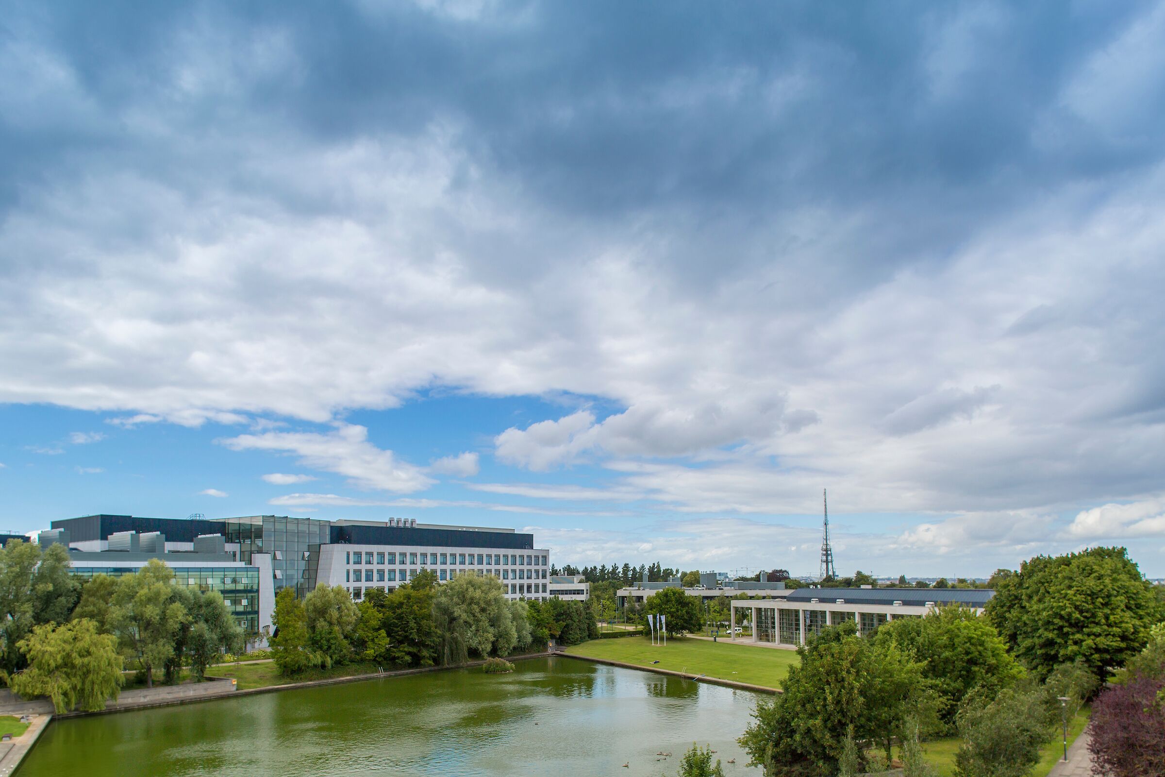 A modern building complex with a large pond, surrounded by trees and greenery.