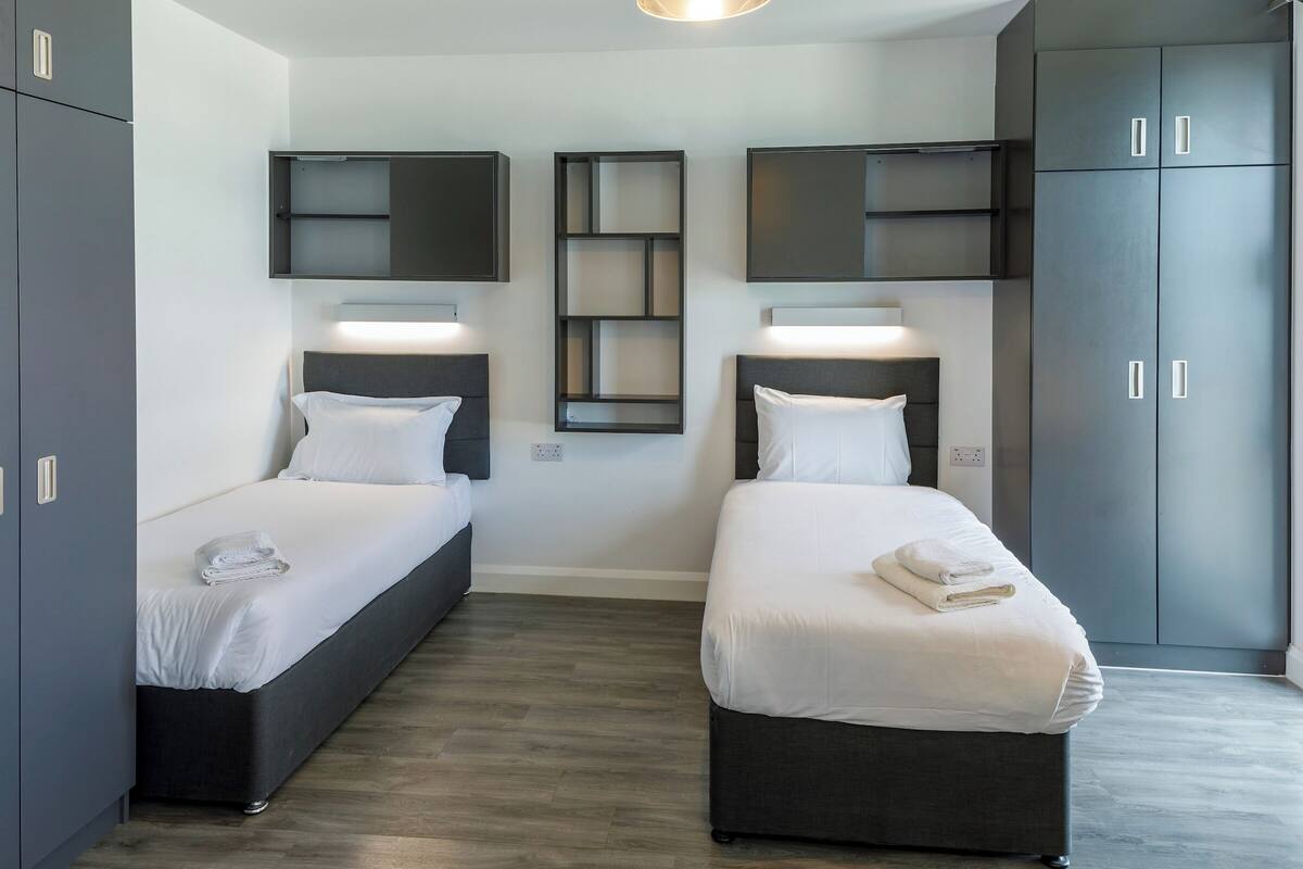 Une chambre d’hôtel moderne avec deux lits simples, des armoires gris foncé et des étagères intégrées au mur.