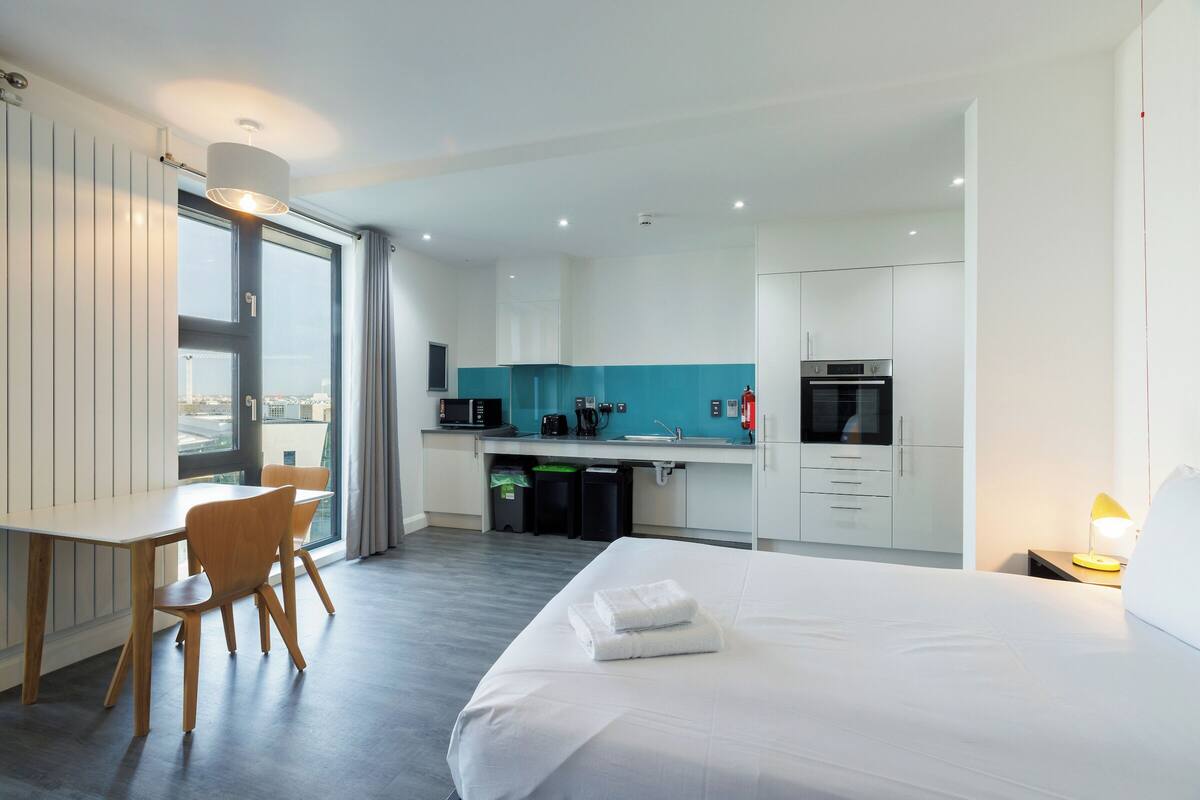 Une chambre d’hôtel moderne dotée d’un grand lit, d’un bureau, d’une chaise, d’une kitchenette et offrant une vue sur la ville.