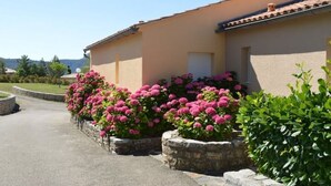 Un bâtiment à la façade beige et au toit recouvert de tuiles rouges. Devant le bâtiment, on peut voir des plantes à fleurs roses.