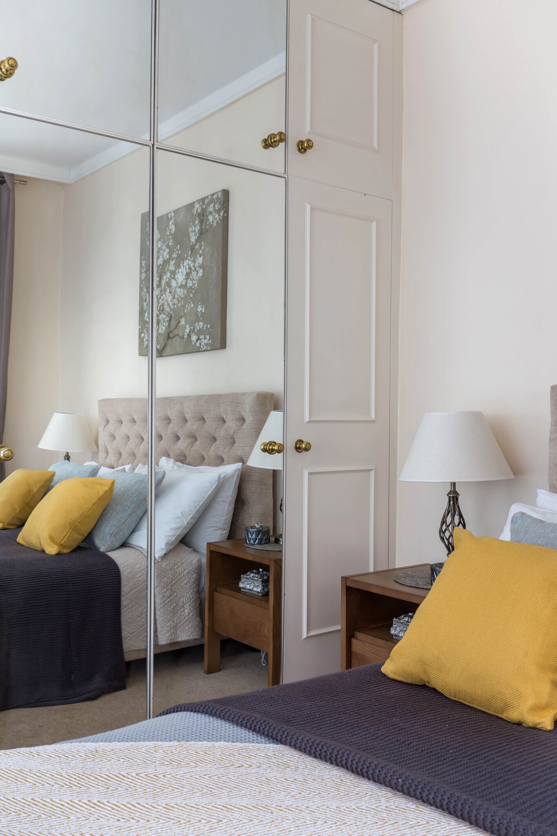 A hotel room with a large mirror, a bed with multiple pillows, a bedside table with a lamp, and a wall-mounted artwork.