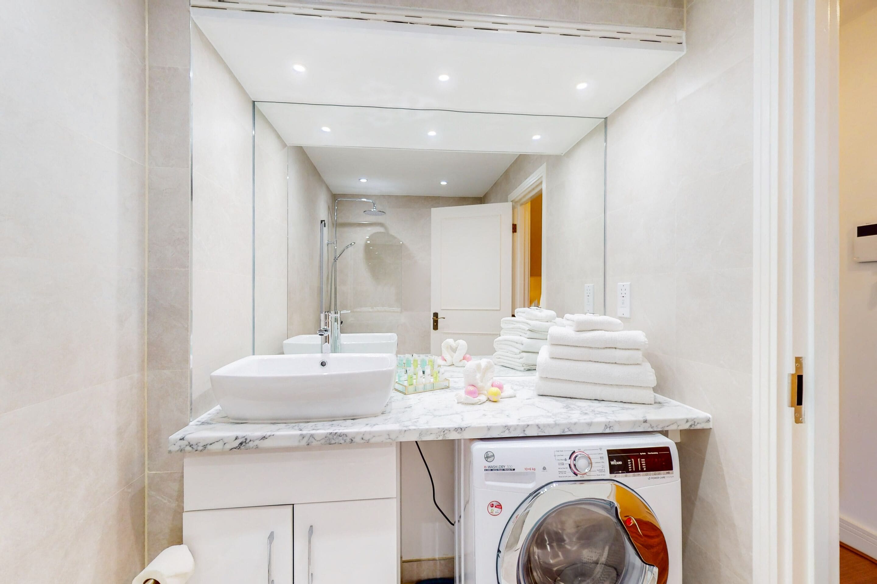A modern bathroom with a marble countertop, a sink, a mirror, a shower, and a washing machine.