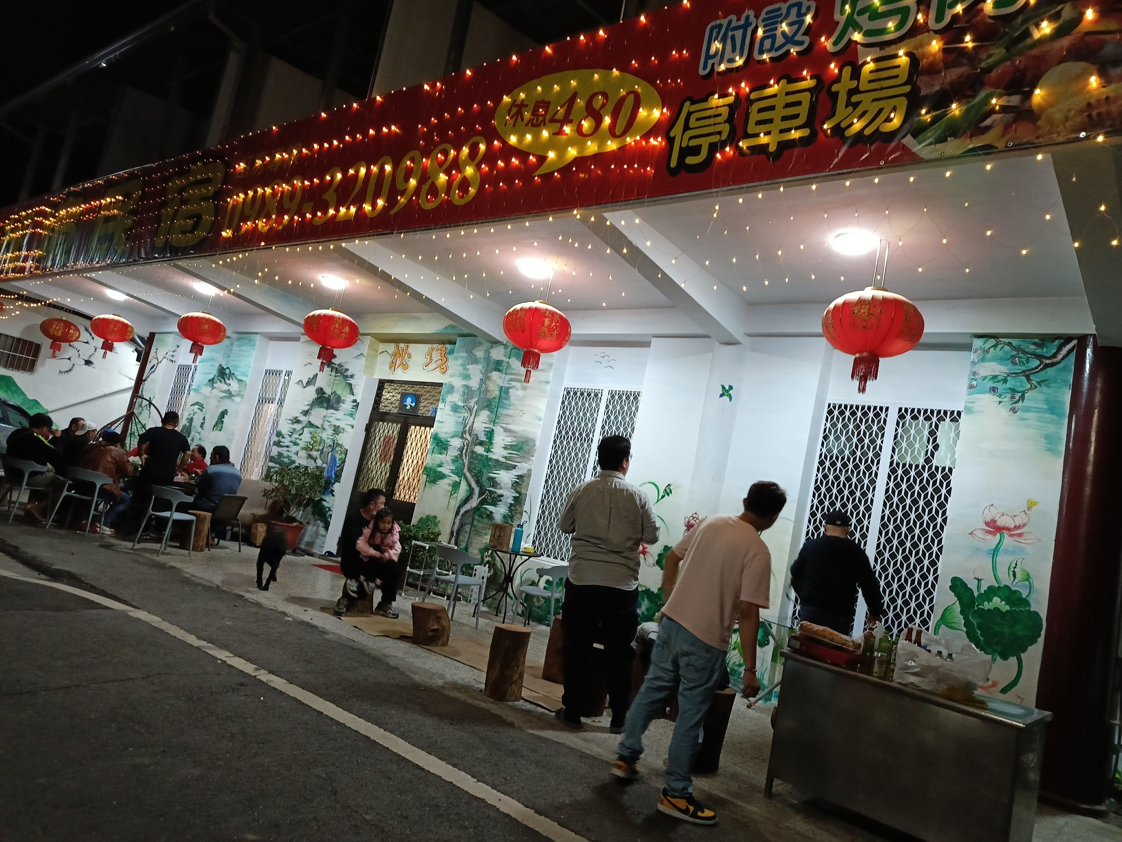 A restaurant with outdoor seating, red lanterns, and a mural of trees.