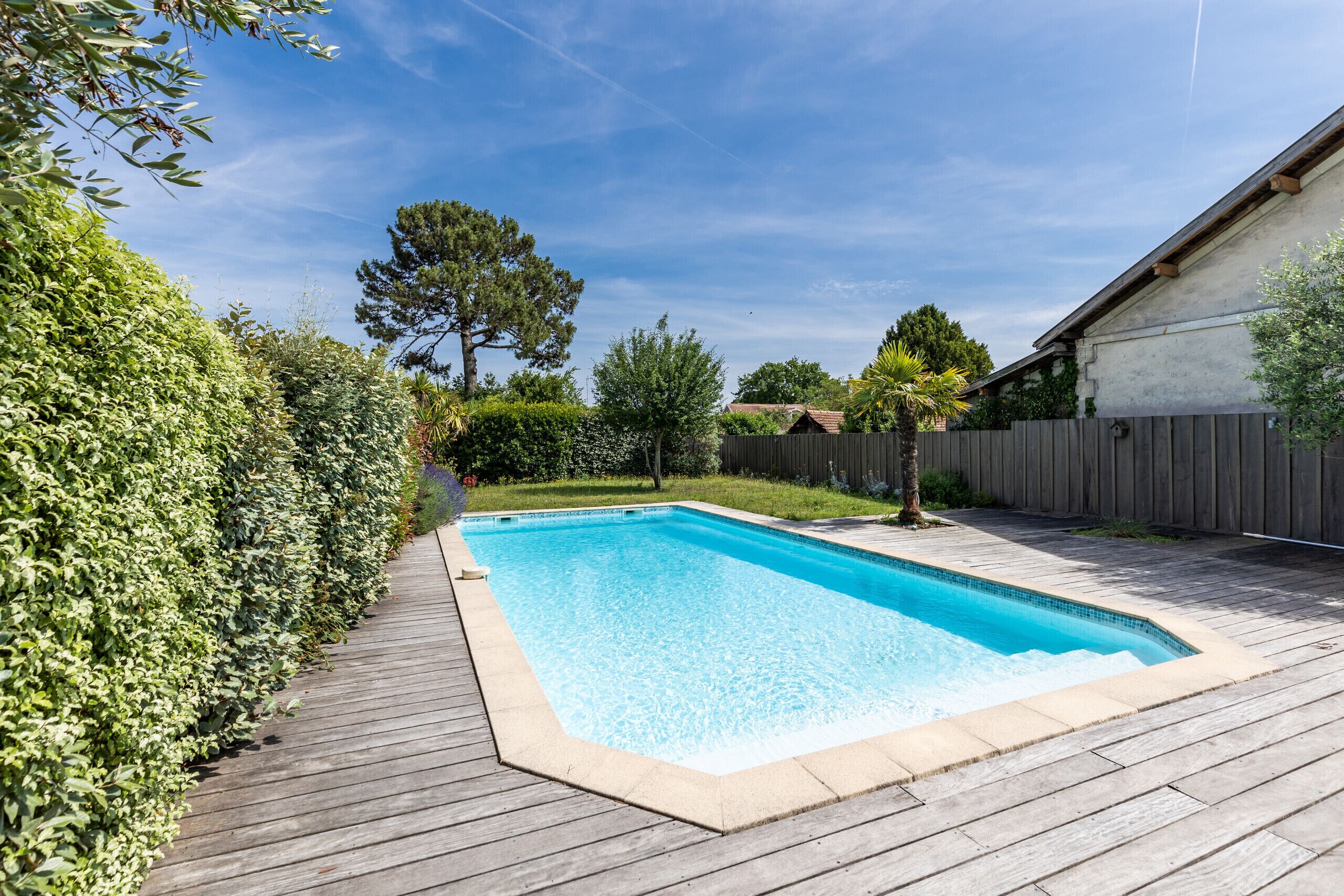 Ein rechteckiger Swimmingpool mit umlaufender Holzterrasse, gelegen in einem Garten mit Holzzaun und verschiedenen Bäumen.