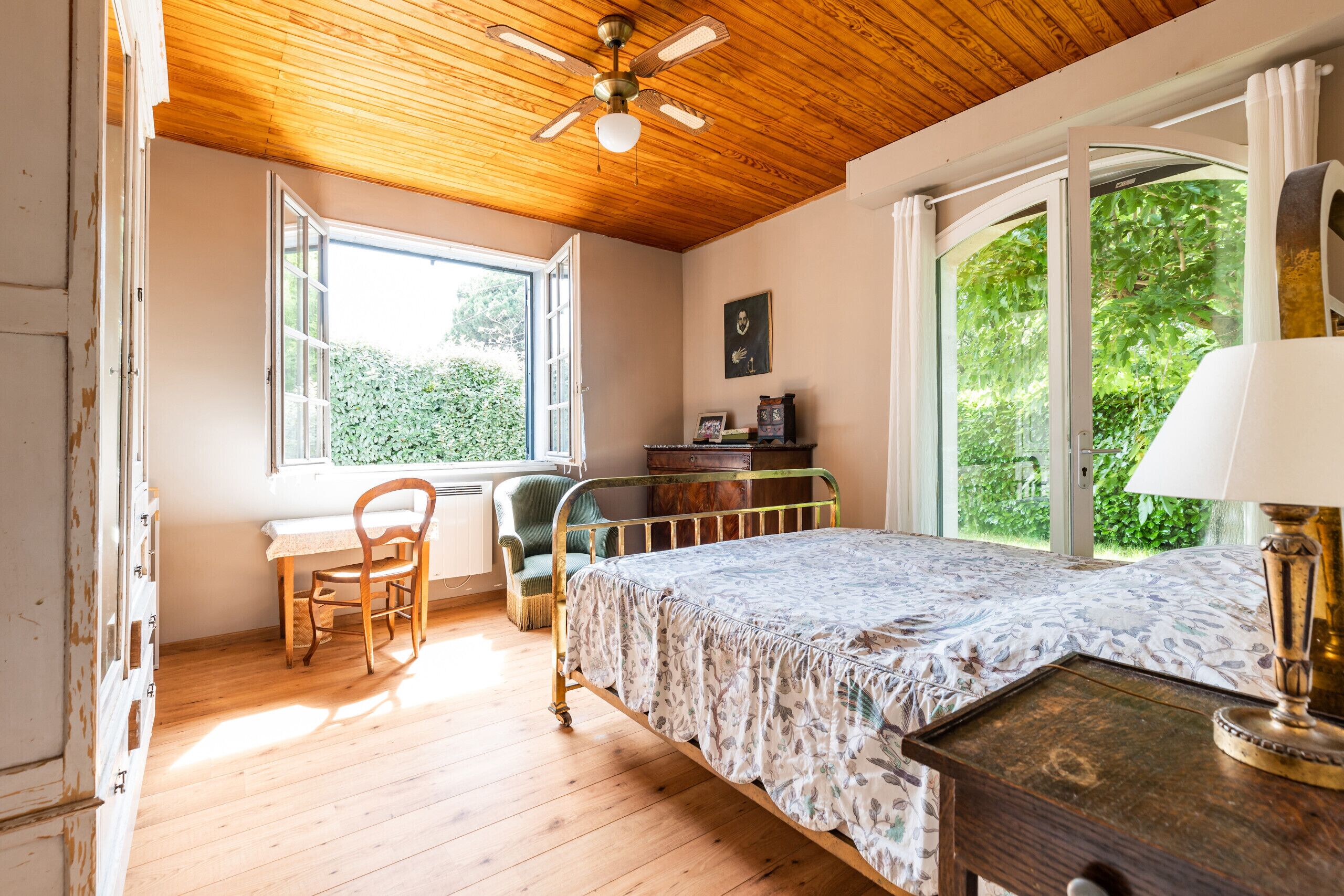 A bedroom with a wooden ceiling, a bed with a floral bedspread, a desk with a chair, and a window with curtains.