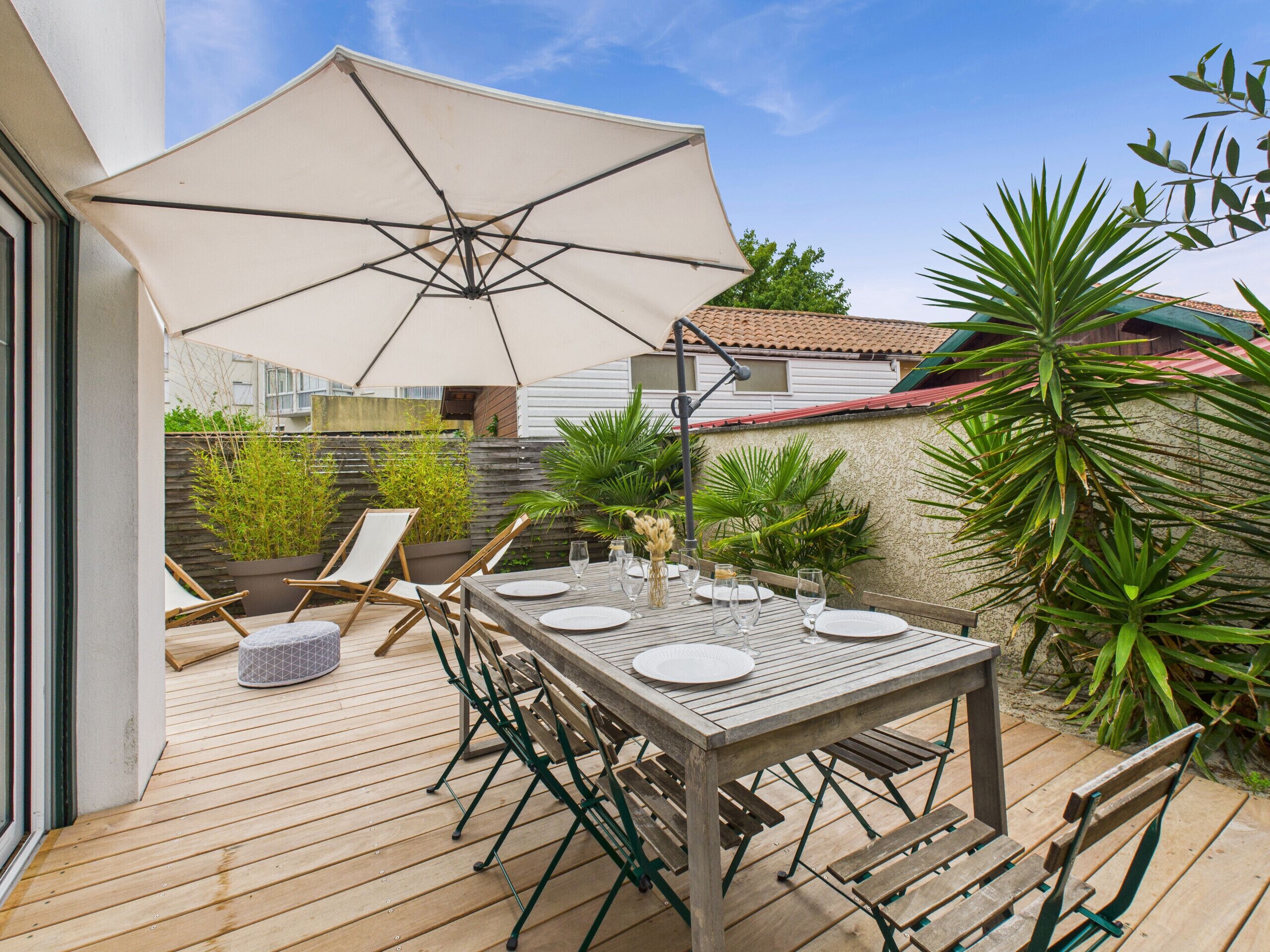 Une terrasse extérieure avec une table en bois dressée pour le repas, entourée de chaises vertes et d’un grand parasol qui offre de l’ombre.