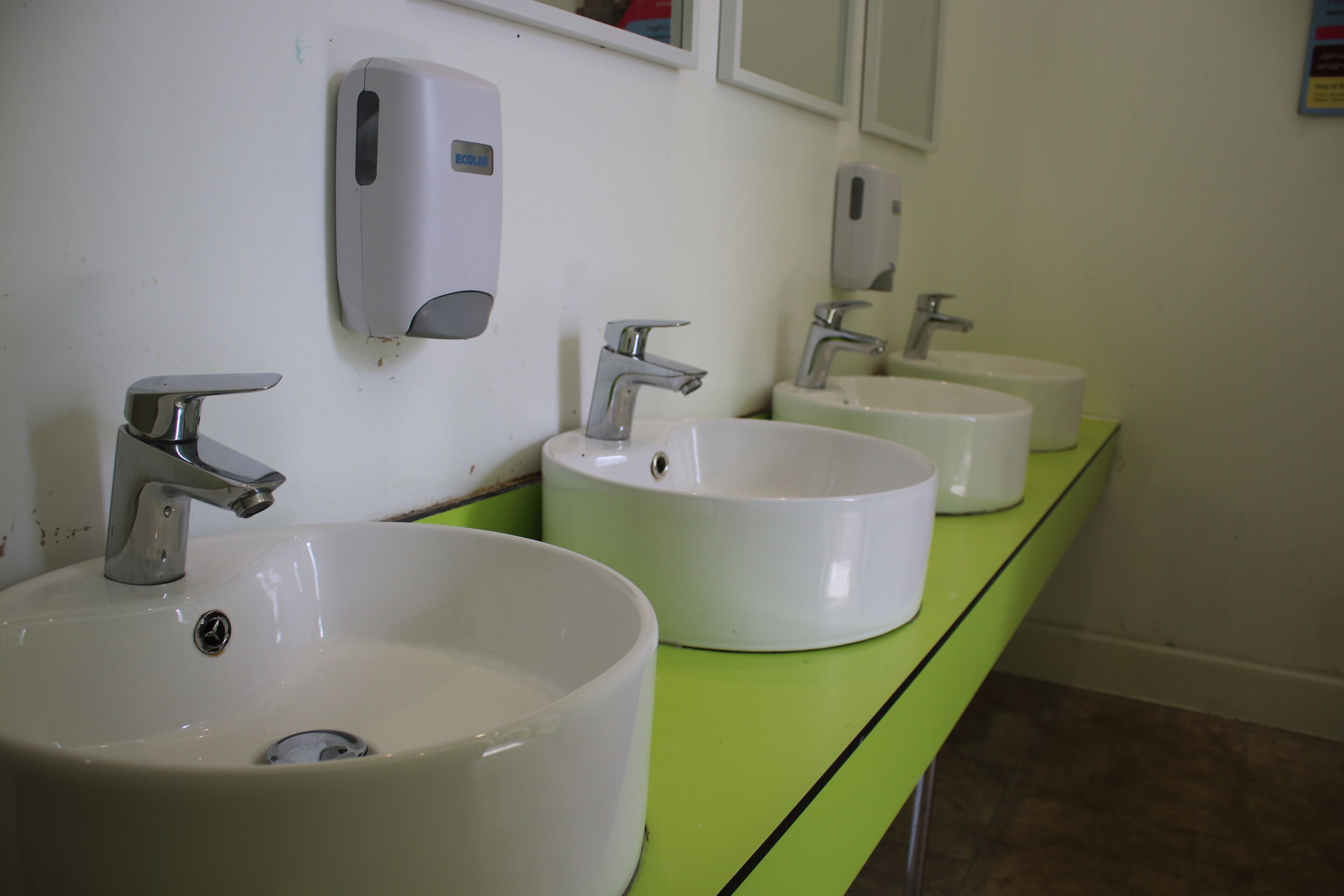 A row of white sinks with chrome faucets and a soap dispenser on a green countertop in a restroom.