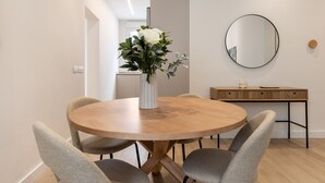 Una mesa de comedor redonda de madera con cuatro sillas grises tapizadas, un jarrón blanco con flores, una consola de madera con un espejo, e interruptor de luz en la pared.