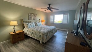 Ein Schlafzimmer mit einem Bett, zwei Nachttischen inklusive Lampen, einem Deckenventilator, einem Fenster mit Jalousien und einem Fernseher auf einem Schrank.