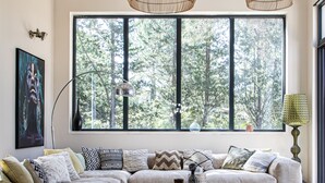 Ein modernes Wohnzimmer mit einer großen Eckcouch, einem Couchtisch und einem großen Fenster mit Blick auf Bäume. Drei hängende Korbtische und eine Stehlampe sind ebenfalls vorhanden.