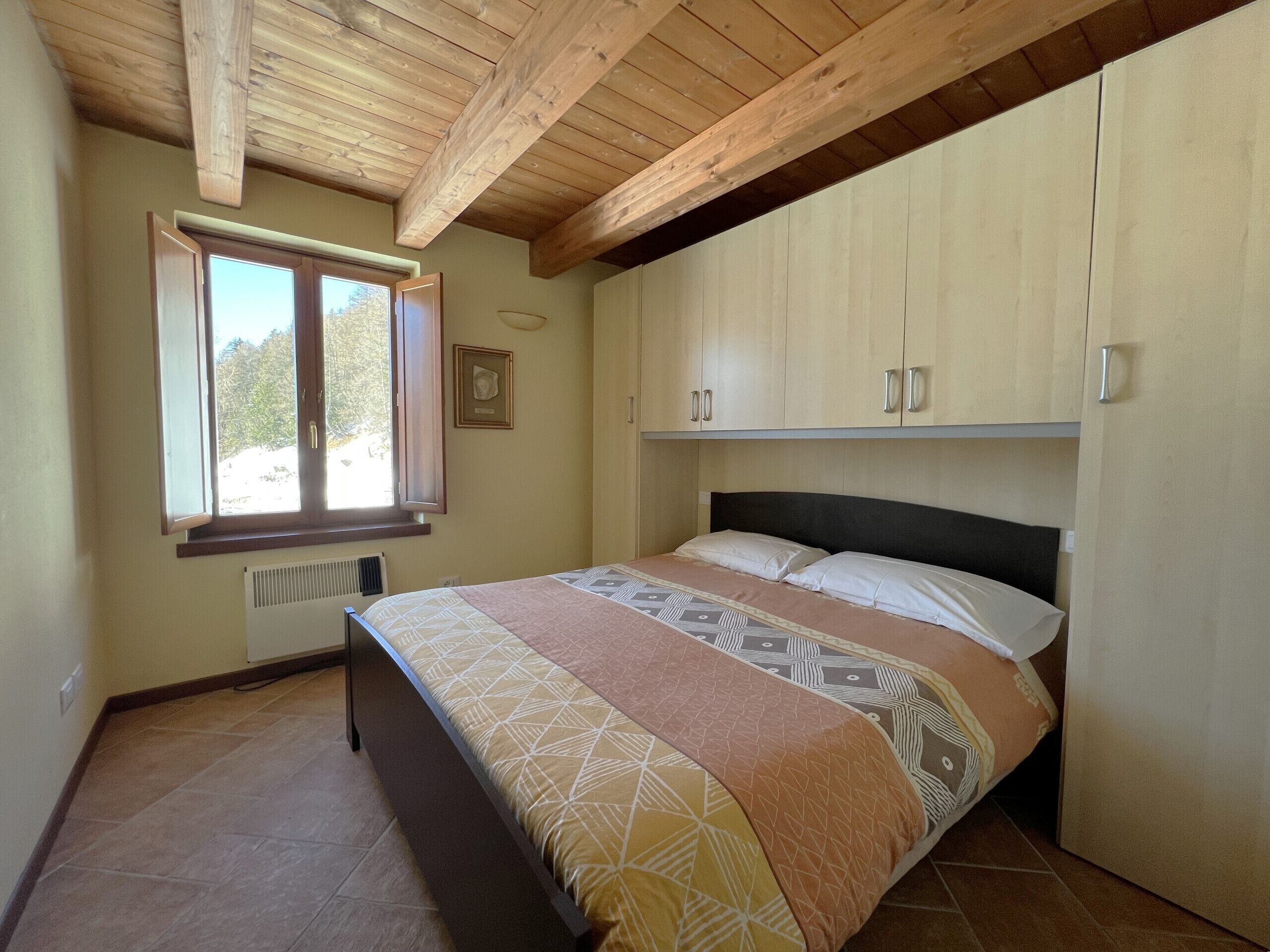 A bedroom with a bed, wooden ceiling beams, a window, and built-in cabinets.