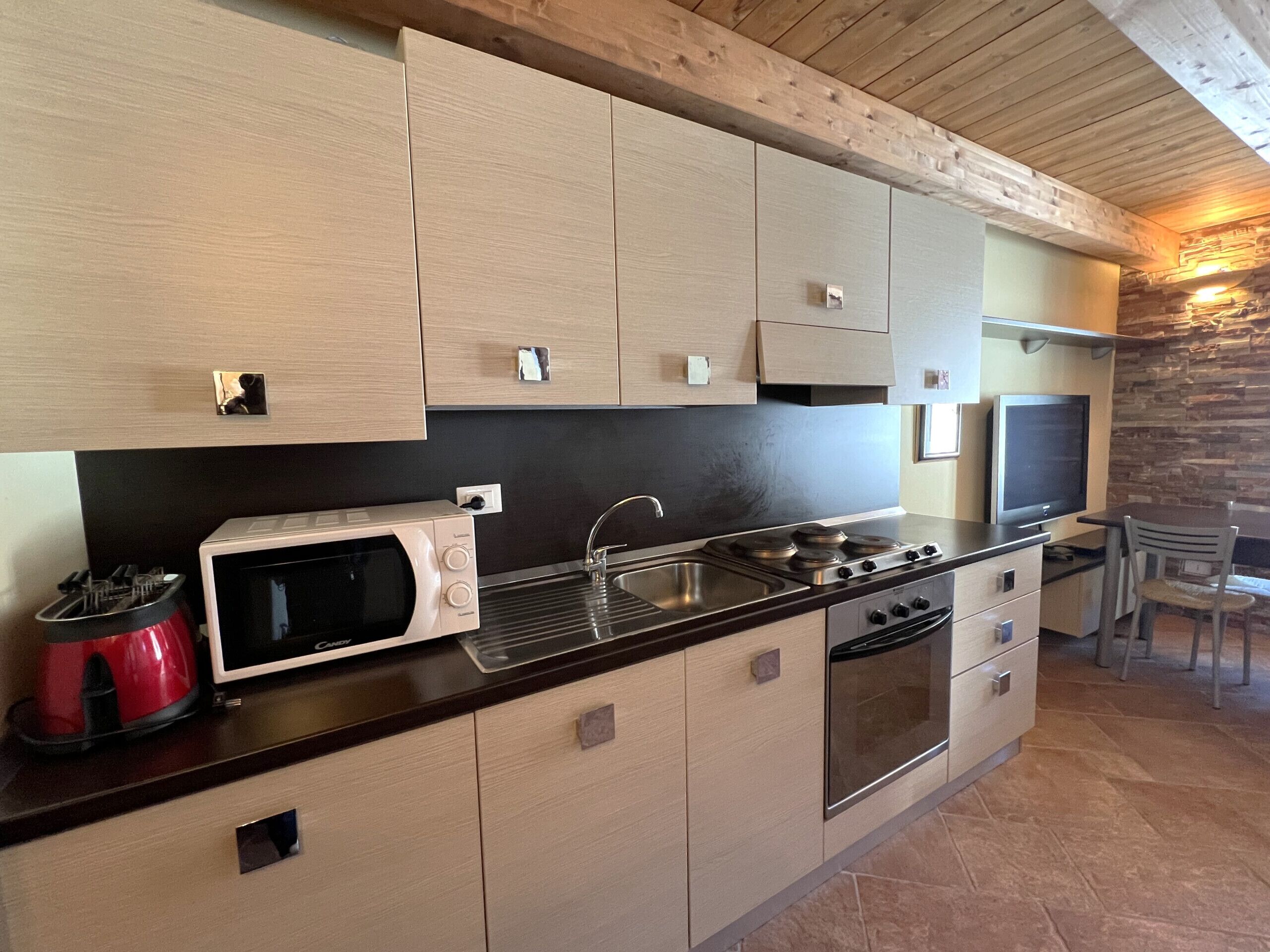 A modern kitchen with light wood cabinets, a microwave, stove, and a dining area with a TV and chairs.