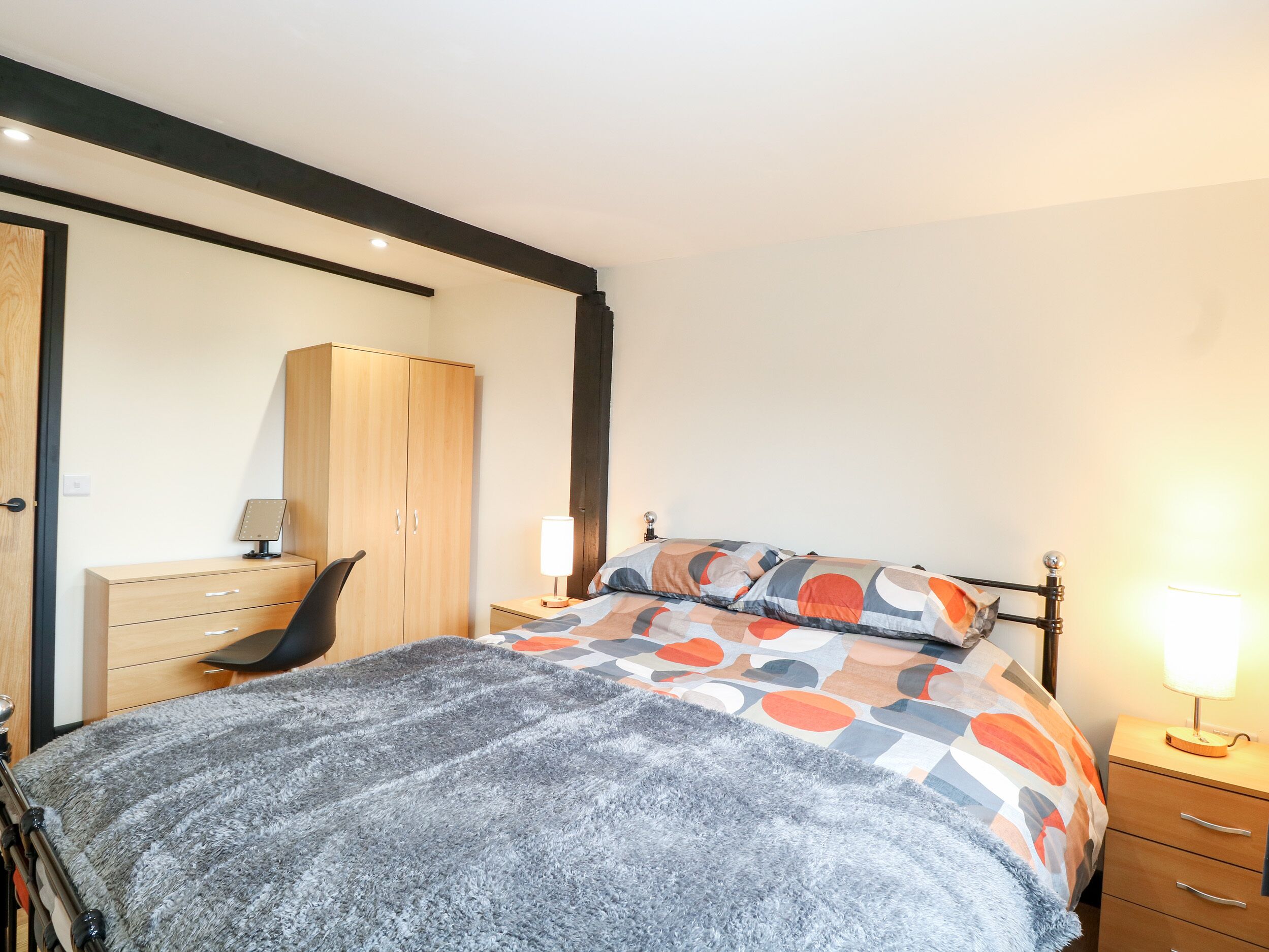 A bedroom with a bed, a wooden wardrobe, a desk with a chair, and a nightstand with a lamp.
