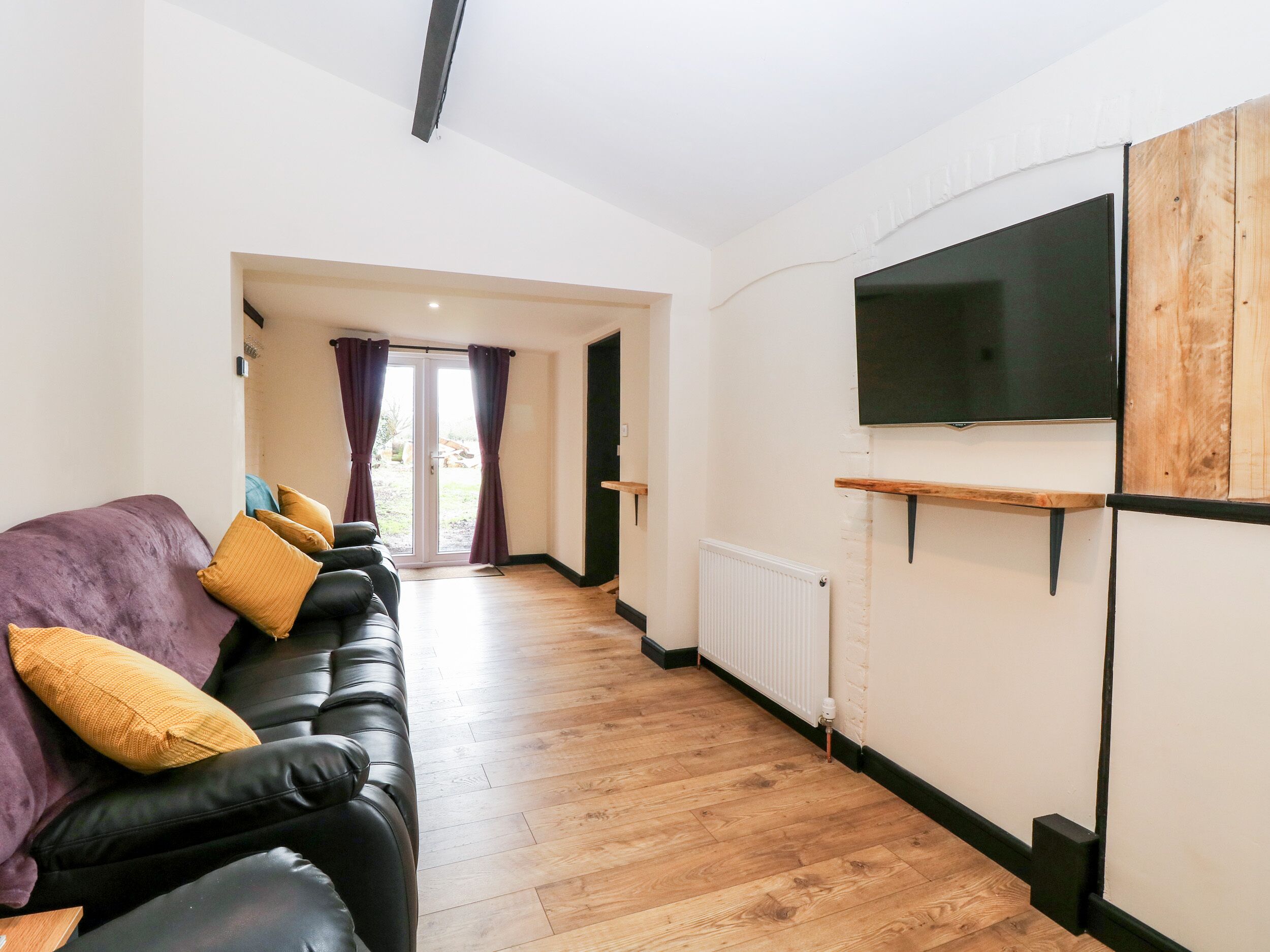 A modern living room with a black leather sofa, wooden flooring, a mounted TV, and a door leading to an outdoor area.