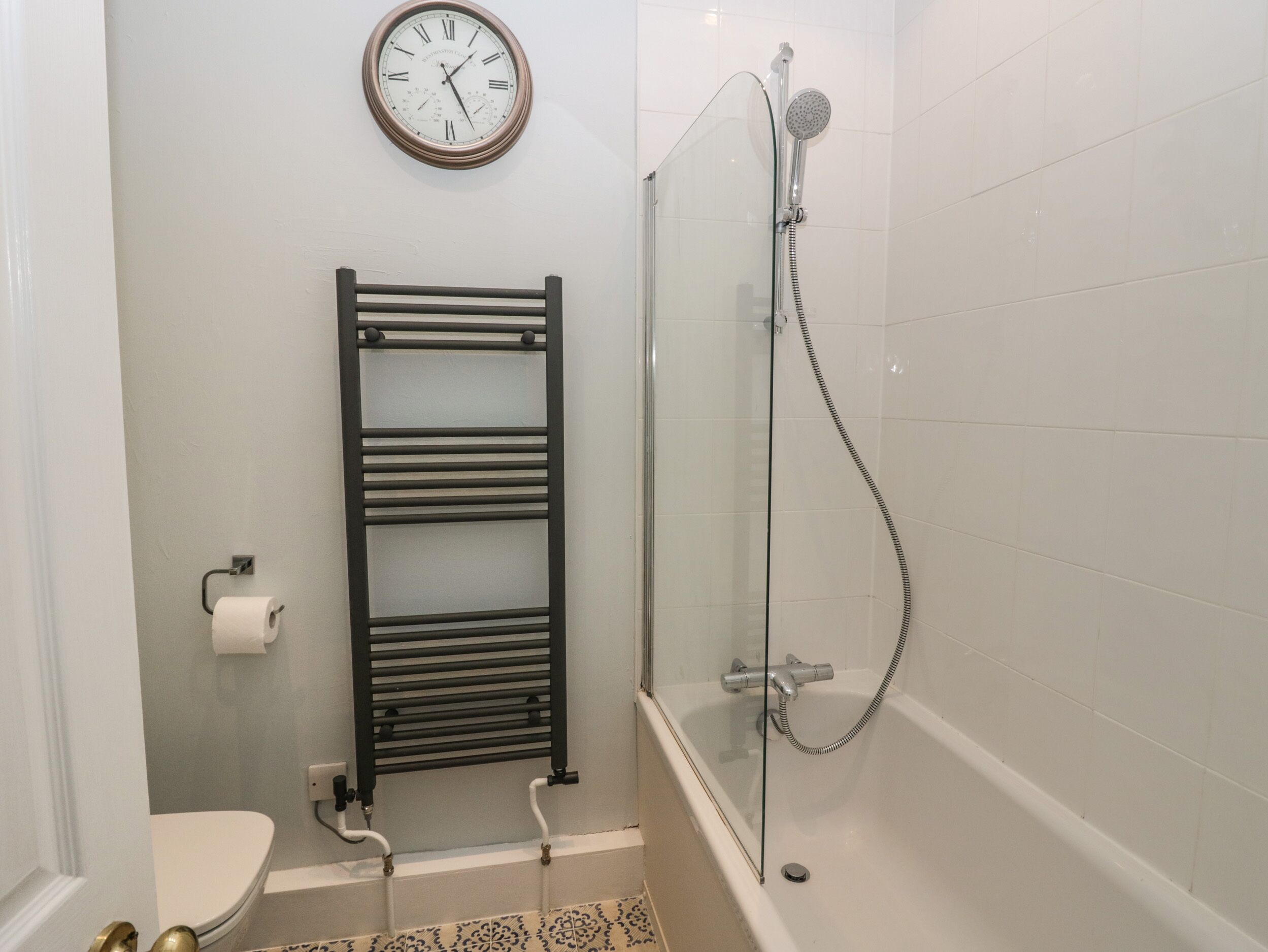 A bathroom with a glass-enclosed shower, a black towel rack, a toilet, and a wall-mounted clock.