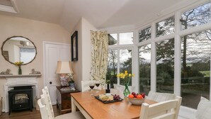 A dining area with a wooden table, white chairs, a fireplace, and a view of the outdoors through large windows. - Summerhow Retreat (Kendal)