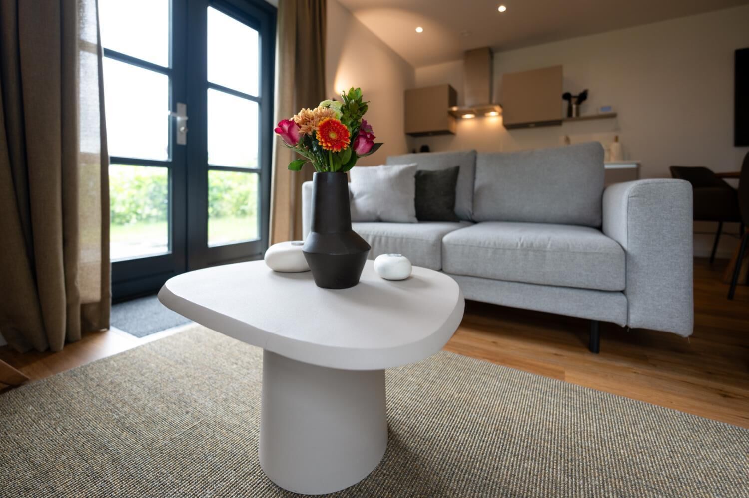 Een moderne woonkamer met een grijze bank, een witte salontafel en een vaas met bloemen.
