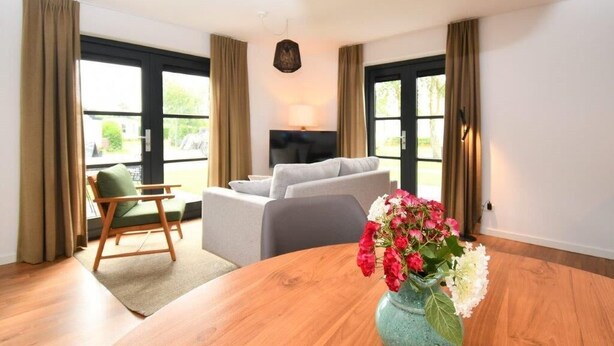 Ein modernes Wohnzimmer mit einem runden Holztisch, einem grauen Sofa und einem grünen Sessel. Ein Fernseher ist an der Wand befestigt und auf dem Tisch steht eine Vase mit Blumen.