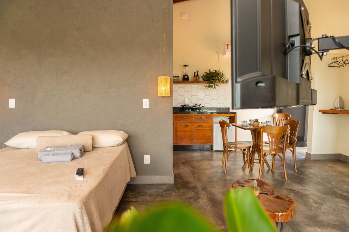 Quarto moderno com uma cama grande, uma mesa de cabeceira e uma pequena mesa de cabeceira com um controle remoto. O quarto possui piso de concreto e teto de madeira.