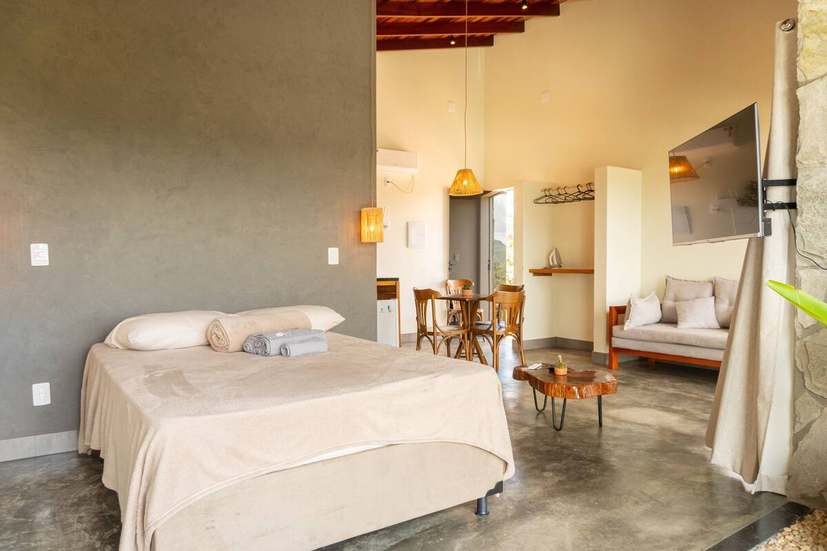 Quarto com uma cama, uma mesinha e uma cadeira. Dá para ver uma área de refeições com uma mesa e cadeiras ao fundo.